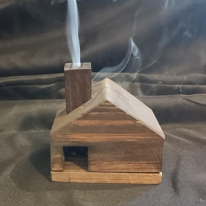 May include: A small, brown wooden house-shaped incense burner with a rectangular chimney. A thin trail of white smoke rises vertically from the top of the chimney. The structure features a gabled roof and a small square opening on the front side.