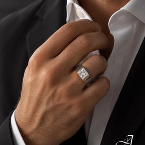 May include: A silver-toned ring featuring a square-cut clear gemstone set in a wide, polished metal band. The ring is worn on a finger, showcasing the faceted stone's brilliance and the smooth, solid construction of the band's metal setting.
