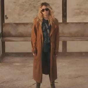 Könnte beinhalten: Ein langer, hellbrauner Trenchcoat über einem dunkelgrauen T-Shirt und einer dunklen Hose. Das Outfit wird mit einer dunklen Sonnenbrille und dunklen Stiefeln kombiniert. Der Mantel hat einen breiten Kragen, lange Ärmel und einen geraden Schnitt.