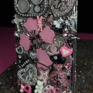 May include: A clear phone case heavily embellished with various charms. The design includes pink crosses, hearts, and the word "Best" in a cow print font. Other charms include a pink bunny, a teddy bear, and a skull.