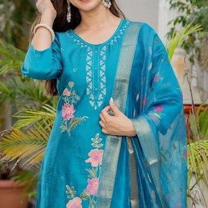 May include: A bright teal kurta featuring a round neckline with silver embroidery, three-quarter sleeves, and a vertical floral pattern in pink and green. The outfit includes a matching sheer teal dupatta with a metallic silver border and floral motifs.