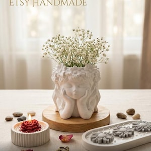 Puede incluir: Una maceta blanca con forma de rostro que contiene flores blancas, sobre una base de madera. Una vela de rosa roja está en un recipiente blanco y acanalado. La imagen también incluye una bandeja decorativa con jabones con forma de flor y el texto "ETSY HANDMADE".