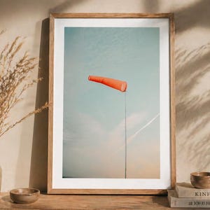 May include: A framed wall art print featuring a bright orange windsock mounted on a thin pole against a pale blue sky with soft clouds. The artwork is displayed in a light wood frame, capturing a clear view of the windsock pointing to the right.