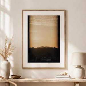 Peut inclure: Une impression d'art mural verticale encadrée représentant un paysage avec un ciel de coucher de soleil doré au-dessus d'une silhouette sombre de collines et de bâtiments lointains, dans un cadre en bois clair avec un large passe-partout blanc.
