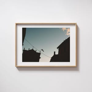 Peut inclure: Photographie encadrée présentant des silhouettes de bâtiments contre un ciel bleu. Un oiseau est en vol, et un fil s'étend entre les bâtiments. Le cadre est en bois clair, et la photographie est montée sur un mur blanc.