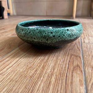 Puede incluir: Un cuenco de cerámica con un borde verde moteado y texturizado y un interior oscuro y brillante. El cuenco se encuentra sobre una superficie de madera, mostrando sus tonos terrosos y su apariencia artesanal. El diseño del cuenco sugiere que podría usarse para servir o exhibir.