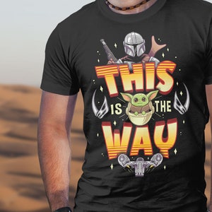 Może przedstawiać: Czarny T-shirt z nadrukiem graficznym postaci Mandalorianina i dziecka Yody. Tekst "This is the Way" jest nadrukowany na koszulce na ż&oacute;łto i pomarańczowo.