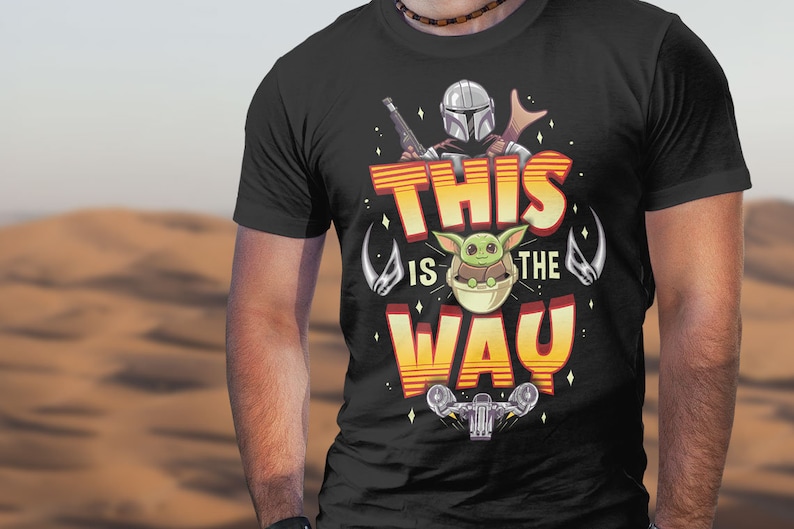 Może przedstawiać: Czarny T-shirt z nadrukiem graficznym postaci Mandalorianina i dziecka Yody. Tekst "This is the Way" jest nadrukowany na koszulce na ż&oacute;łto i pomarańczowo.