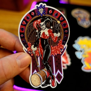 May include: A sticker featuring a colorful illustration of Harley Quinn, a comic book character, holding a mallet. She is dressed in a red and black costume with a jester's hat. The background is a dark red and black design with playing card symbols.