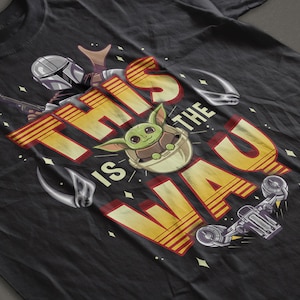 Może przedstawiać: Czarny T-shirt z ż&oacute;łto-czerwonym nadrukiem przedstawiającym tekst "This is the Way" z kresk&oacute;wkową postacią zielonego kosmity i postacią Mandalorianina.