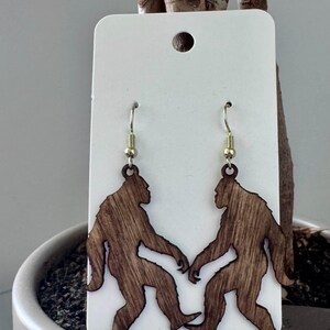May include: Wooden earrings featuring a Bigfoot design. The earrings are cut from wood and stained a medium brown color. The Bigfoot figures are holding hands. The earrings hang from gold-colored fish hook ear wires.