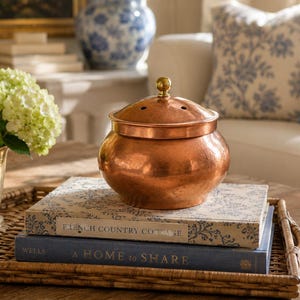 Puede incluir: Una olla redonda de cobre martillado con una tapa a juego que presenta un pequeño pomo de latón y orificios de ventilación. La olla descansa sobre dos libros apilados, titulados French Country Cottage y A Home to Share, en una bandeja tejida.