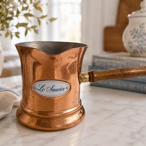 Peut inclure: Une casserole en cuivre avec un corps cylindrique, un bord évasé et un long manche en bois. Une plaque ovale en émail blanc sur le devant porte le texte bleu Le Saucier. La casserole a une finition métallique et une base large.