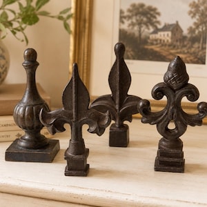 Pode incluir: Um conjunto de quatro remates decorativos castanho-escuros com formas variadas, incluindo uma peça arredondada estilo urna, dois designs pontiagudos de flor-de-lis e uma peça ornamentada com volutas e topo texturizado, todos com bases quadradas.