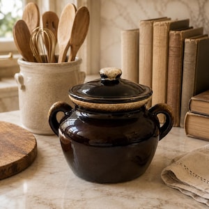 May include: A dark brown glazed ceramic bean pot with two side handles and a matching lid. The pot features a lighter tan rim and a rounded body shape. It is a functional kitchen vessel designed for cooking or storage, showcasing a glossy finish.