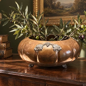 May include: A round, hammered copper bowl featuring silver-toned metal handles shaped like grape clusters and leaves. The bowl rests on three small, decorative silver-toned feet and is filled with green olive branches with small dark olives.