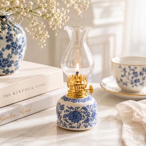 Peut inclure: Une petite lampe à huile avec une base en céramique bleue et blanche à motifs floraux, un brûleur en métal doré et une cheminée en verre transparent. La lampe présente une mèche allumée, créant une lueur chaude sur la base décorée.