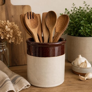 May include: A ceramic utensil holder with a brown band and off-white base, filled with wooden spoons and a fork. The kitchen scene includes a wooden cutting board, a small bowl of garlic, and a potted plant.