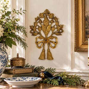 May include: A gold-colored wall plaque featuring a symmetrical floral arrangement with leaves and flowers, tied at the bottom with a decorative bow and two hanging tassels. The piece has a textured, metallic finish and is mounted on a wall.