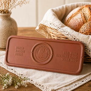 May include: A terracotta-coloured bread basket brick with a rectangular shape and rounded edges. The brick features embossed text, including "Bread Basket Brick" and "Use with Extreme Care When Hot." A logo is in the center. A basket of bread is in the background.