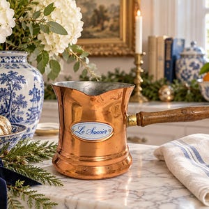 May include: A copper sauce pot with a long wooden handle and a pouring spout. The pot features a white oval enamel plaque on the front with the blue script text Le Saucier. The metal surface shows a polished finish with minor signs of wear.