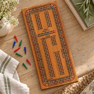 May include: A rectangular wooden cribbage board featuring a printed track with black, red, and blue holes. The board is labeled Club Cribbage Master, Drueke Model 1977, and includes several small plastic pegs in red, blue, and green colors.