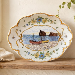 May include: An oval ceramic platter with a scalloped edge, featuring a hand-painted coastal scene of two small boats on a beach with rock formations in the sea. The border is decorated with yellow ribbons, blue and yellow flowers, and small insects.