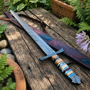 May include: A long, ornate sword with a patterned blade and a multi-colored handle rests on a weathered wooden surface. The sword has a cross-shaped hilt and is accompanied by a brown leather sheath with a shoulder strap. The blade has a length of approximately 28 inches.