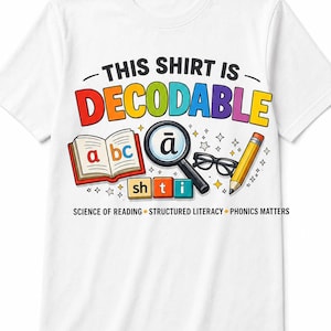 May include: A white t-shirt featuring a colorful graphic with the text THIS SHIRT IS DECODABLE. The design includes an open book with letters a, b, c, a magnifying glass over the letter ā, black glasses, a yellow pencil, and blocks with sh, t, i.