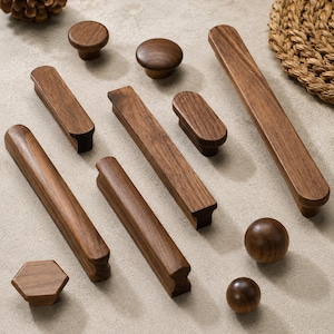 May include: A collection of various wooden cabinet hardware, including knobs, pulls, and a hexagonal knob. The hardware is made of dark brown wood and arranged on a neutral surface. A pinecone and woven placemat are also present.