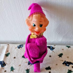 May include: A vintage-style elf figurine featuring a soft, bright pink felt body and a matching pointed hat. The elf has a moulded plastic face with painted blue eyes, rosy cheeks, and a red mouth, accented by a small, shiny gold metallic collar.