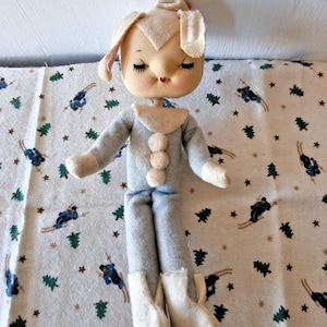 May include: A plush doll with a cream-coloured face, closed eyes, and a small red heart-shaped mouth. The doll wears a light blue one-piece suit with three round white buttons down the front, a cream-coloured collar, and matching cream-coloured boots.