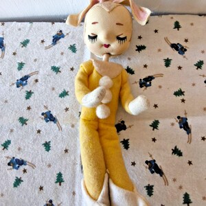May include: A vintage-style plush doll with a cream-colored head, long floppy ears, and closed eyes with long eyelashes. The doll wears a yellow long-sleeved jumpsuit featuring three white pom-poms down the front, white cuffs, and white booties.