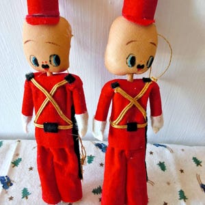 May include: Two vintage-style toy soldier ornaments featuring fabric heads with painted facial features, red felt uniforms, gold braided trim, black belts, and tall red cylindrical hats. Each figure has white cuffs and a gold hanging cord attached.