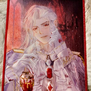 May include: A framed print featuring a detailed illustration of a person with long white hair, wearing a white and lavender outfit with gold accents. The artwork has a gothic theme, with a red and gold color scheme.