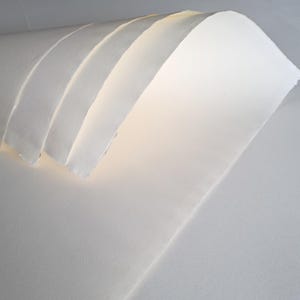 May include: A stack of several sheets of thick, white, textured paper with uneven, deckled edges. The sheets are curved and layered, creating a soft, flowing appearance with subtle shadows between each individual layer of paper.