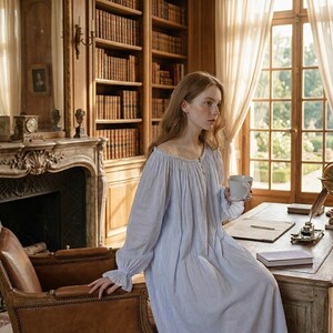 May include: A light blue nightgown with long sleeves and a ruffled neckline. The nightgown is buttoned down the front and is worn by a person holding a white mug. The scene is set in a room with a fireplace, bookshelves, and a desk.