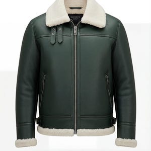 Peut inclure: Veste en cuir vert foncé avec un col et une doublure en peau de mouton crème. La veste est dotée d'une fermeture éclair, de deux poches latérales et de boucles. La marque "MAVERICK JACKET" est visible sur le col.