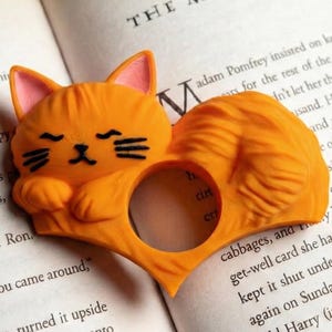 Op de afbeelding: Een oranje katvormige boekenlegger met een hartvormige ring. De kat heeft gesloten ogen en een gekrulde staart. Het boek is open op een pagina met de titel "THE MARAUDER'S MAP".