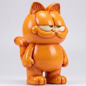 Puede incluir: Una figura de gato naranja de pie con cabeza redonda, orejas pequeñas y una expresión facial de suficiencia. El personaje tiene un cuerpo regordete, extremidades cortas y una cola rayada visible en el lado izquierdo. La superficie es lisa y brillante.