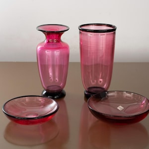 May include: A collection of four pink and black glass items. The set comprises a vase, a tall glass, and two small bowls. The vase and tall glass feature black rims and bases. The bowls also have black rims.