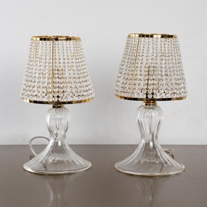 May include: Two clear glass table lamps with crystal bead shades and gold trim. The lamps have a classic design with a clear glass base and a decorative shape. The shades are made of small, clear crystals.