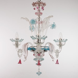 Op de afbeelding: Een heldere glazen kroonluchter met roze en blauwe bloemaccenten. De kroonluchter heeft drie armen met lampen en decoratieve glazen elementen. Het ontwerp omvat hangende roze glazen hangers en een centrale lichtcluster.