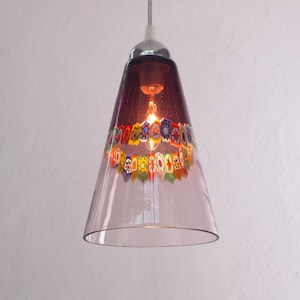 Può includere: Una lampada a sospensione con un paralume conico in vetro viola trasparente. Il paralume presenta una fascia decorativa di motivi floreali colorati vicino alla parte superiore. La lampada ha una parte superiore cromata e una singola lampadina.