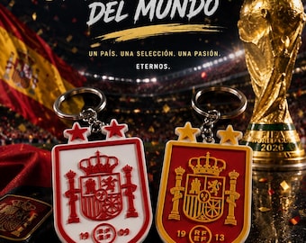 May include: Two shield-shaped keychains featuring the Spanish coat of arms and the RFEF logo. One is white with red details, the other is red with yellow details. Both have two stars on top. The background displays the 2026 World Cup trophy.
