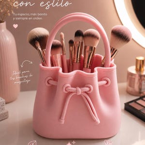 Puede incluir: Un porta brochas de maquillaje de silicona en color rosa claro con forma de cubo, asa superior y un detalle de cordón decorativo. El recipiente contiene varias brochas de maquillaje con virolas de oro rosa y cerdas suaves.