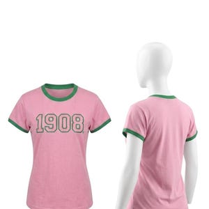 May include: A pink ringer t-shirt with green trim on the crew neckline and sleeve cuffs. The front of the shirt features the number 1908 printed in a green font with a dark outline. The shirt is shown from both the front and back views.