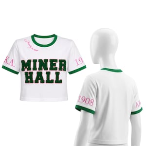 Könnte beinhalten: Ein weißes bauchfreies T-Shirt mit grünem Saum an Kragen und Ärmeln. Die Vorderseite zeigt den Text MINER HALL in grünen Chenille-Buchstaben sowie rosa Text mit January 15, 1908, A.K.A. und 1908 auf den Ärmeln und am Halsausschnitt.