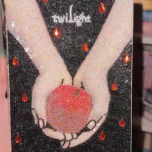 May include: A decorative book cover featuring the word "Twilight" in white script. The cover depicts two hands holding a red apple, surrounded by black and white beads and red teardrop-shaped accents. The overall design is embellished with sparkling details.