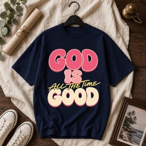 Pode incluir: Uma t-shirt azul-marinho de manga curta com o texto "GOD IS GOOD" em letras grandes e arredondadas cor-de-rosa e creme, com a frase "ALL THE TIME" escrita em letra amarela entre as palavras "IS" e "GOOD" na parte da frente.